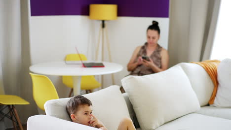child relaxes on couch while adult texts. a young boy sits comfortably on a couch, focused on something ahead, while an adult is busy texting in the background