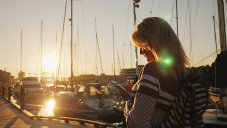 Una-Mujer-Joven-Está-Usando-Un-Teléfono-Inteligente-De-Pie-En-Un-Muelle-Al-Atardecer-En-El-Fondo-Se-Puede-Ver-Belleza