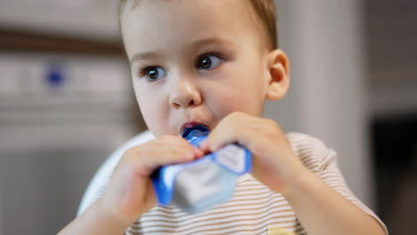 cute caucasian kid eating fruit puree from a doy pack. lovely baby likes healthy food. close up. blurred backdrop.