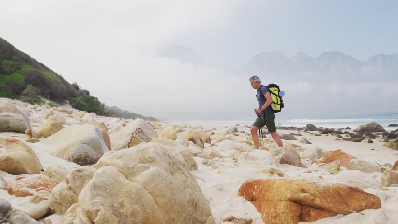hombre excursionista sénior con mochila y palos de senderismo caminando mientras caminaba por la playa.