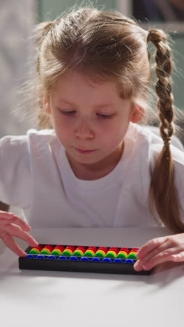 pretty little girl student learns to solve tasks using toy abacus sitting at white desk during arithmetic lesson at home closeup slow motion