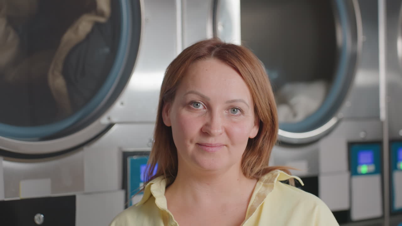 Portrait of elegant woman in casual standing before industrial laundry dryers, clothes rolling inside behind glass, stainless panels, displays, warm smile, modern laundromat, digital controls glowing
