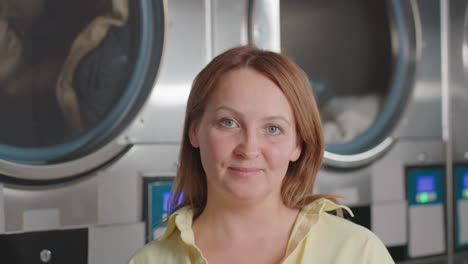 Portrait of elegant woman in casual standing before industrial laundry dryers, clothes rolling inside behind glass, stainless panels, displays, warm smile, modern laundromat, digital controls glowing