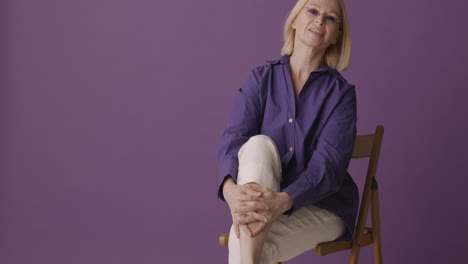 blonde mature woman with purple shirt, beige pants and sunglasses posing looking at camera sitting on chair with crossed legs on purple background