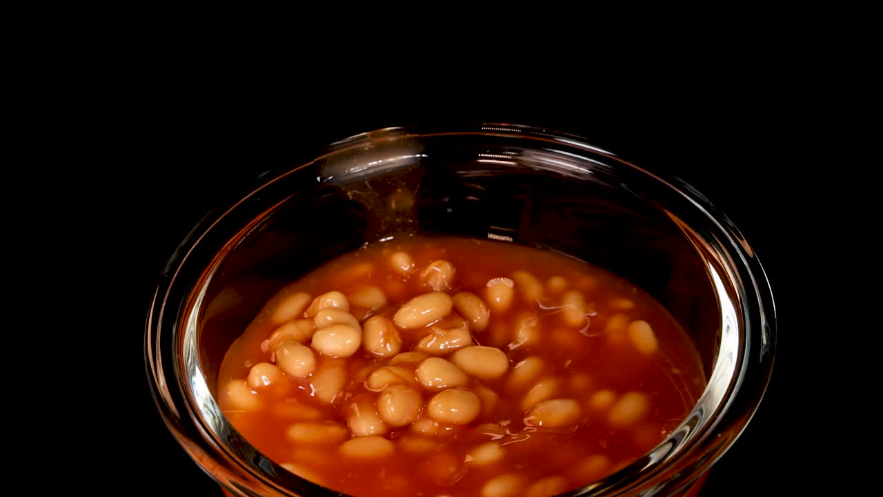 Baked beans in tomato sauce are dropped into a clear glass bowl, captured in slow motion with dramatic studio lighting and a black background