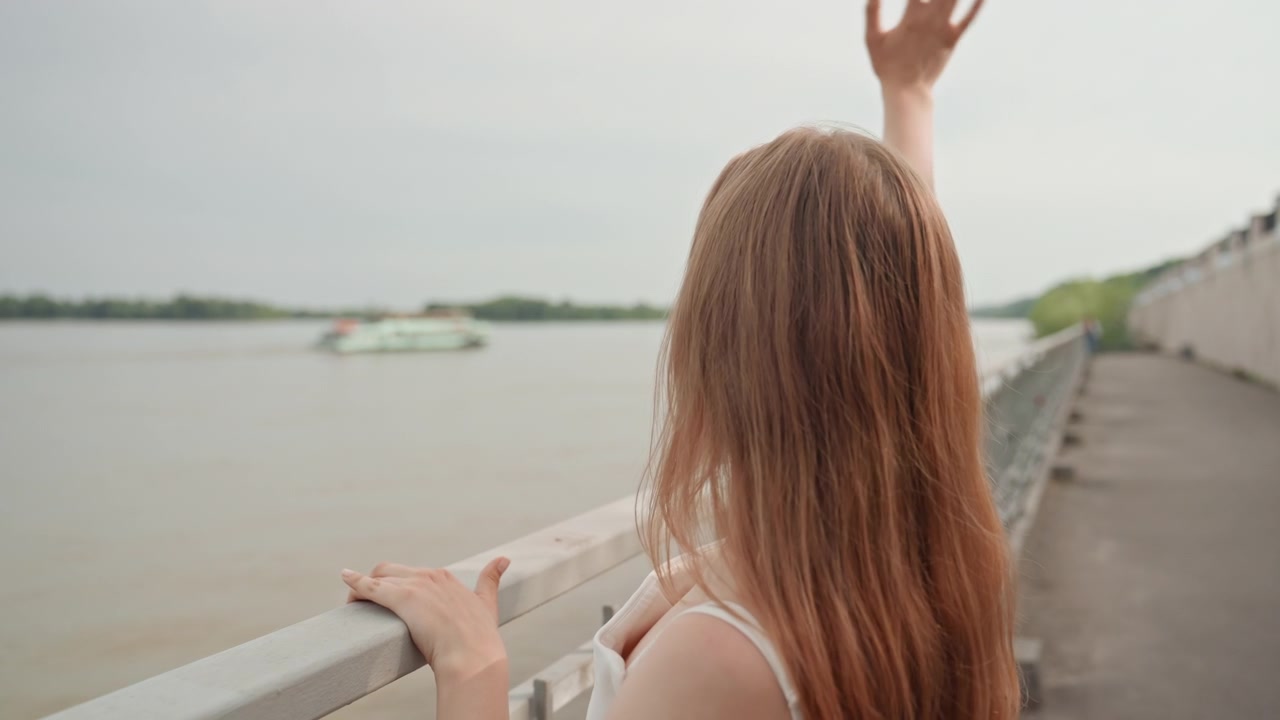 Woman with long hair stands on riverside walkway gazing out at water and distant landscape. Woman rests one hand on metal railing while raising her other arm upward creating sense