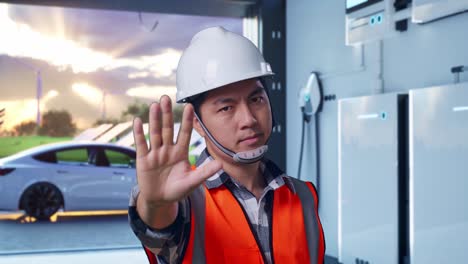 close up of asian male engineer with safety helmet disapproving with no hand sign while standing with home energy storage system in a modern garage