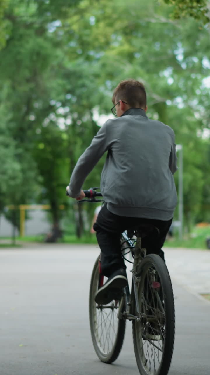 나무로 둘러싸인 푸른 공원을 가로질러 함께 자전거를 타고 다니는 두 친구의 뒷면, 두 사람은 길을 따라 동기화되어 달리고 있으며, 멀리서 걸어가는 사람들의 흐릿한 인물이 있습니다.