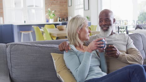 Una-Pareja-De-Ancianos-De-Raza-Mixta-Tomando-Café-Juntos-Mientras-Están-Sentados-En-El-Sofá-De-Casa