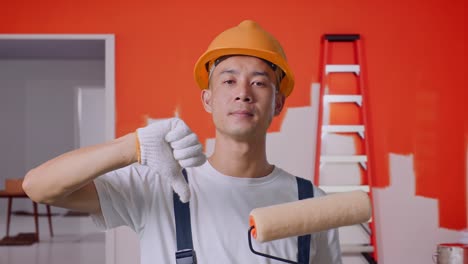 close up of asian man painter wearing safety helmet showing thumbs down gesture to camera while standing with painting wall in room
