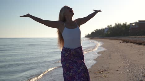 Mit-Blick-Auf-Die-Kamera-Hebt-Eine-Reife-Frau-Im-Gegenlicht-Am-Strand-Ihre-Arme-Und-Blickt-In-Den-Himmel