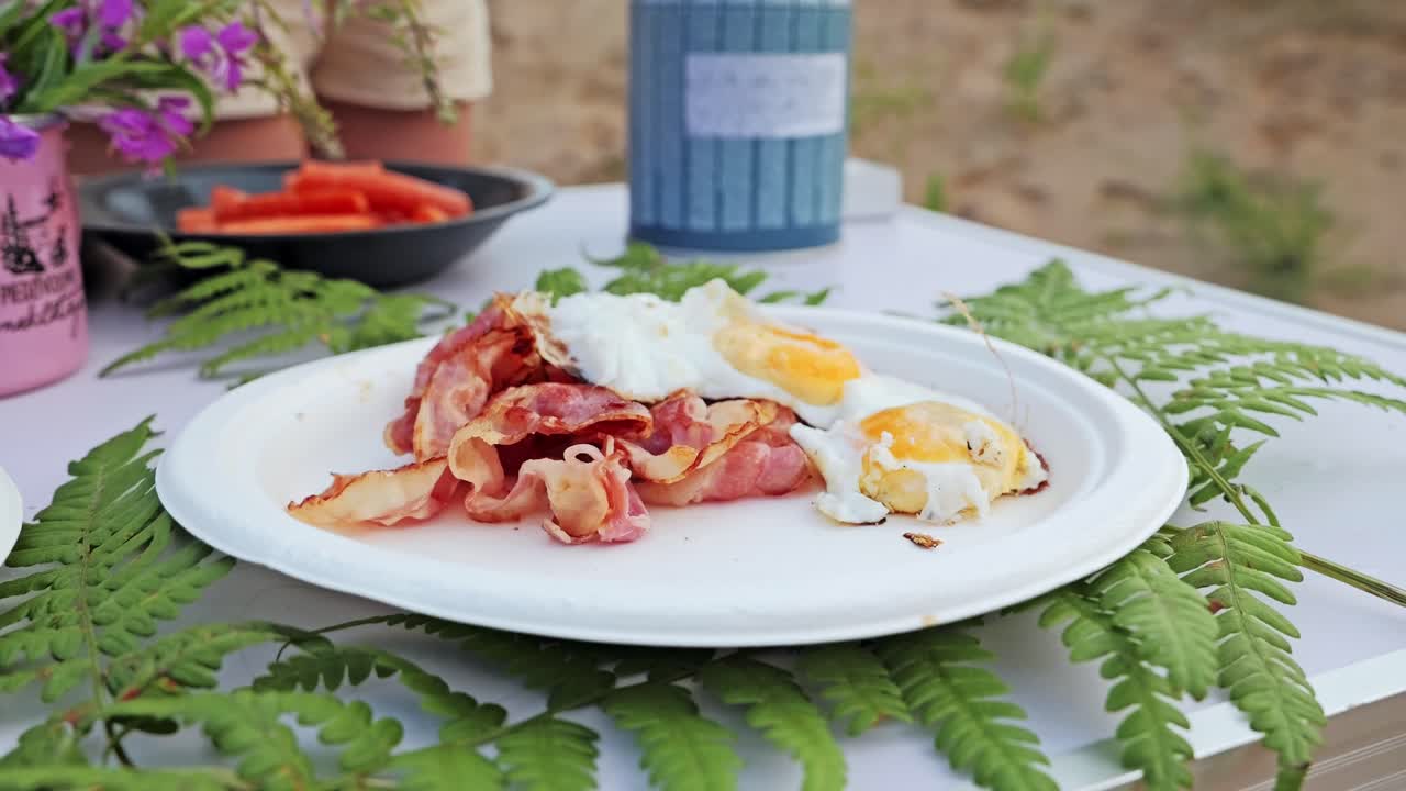 Close up of fried eggs placed onto hot bacon plate on fern-covered picnic table