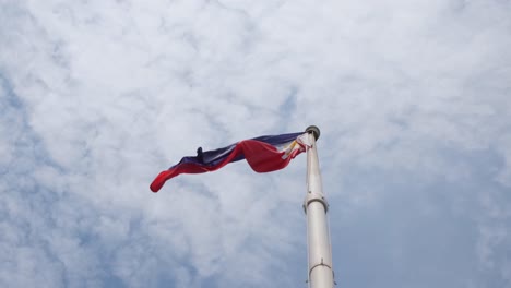 Philippine-National-Flag-flies-towards-the-left-while-the-camera-tilts-up-a-little,-fluffy-clouds-at-the-background