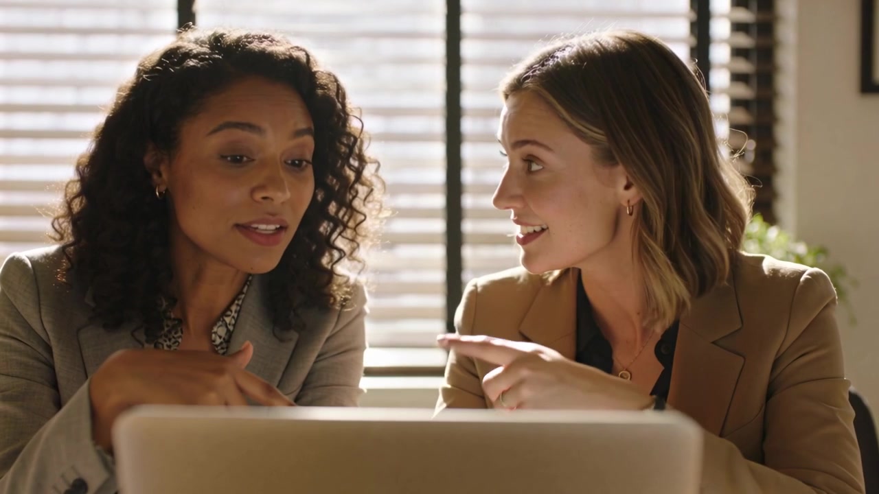 Viewing laptop screen, two women laughing, reviewing content and leaning forward at desk, blazers