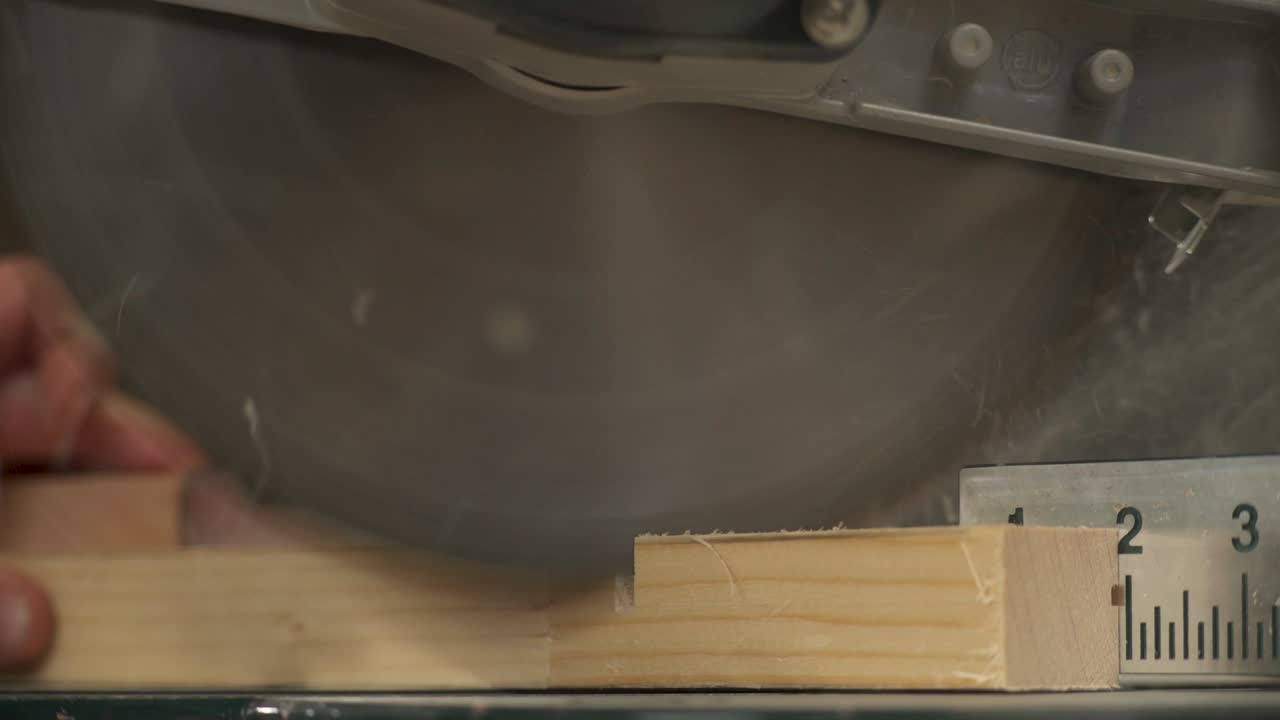A close-up of a miter saw making a cross-cut of a work piece with sawdust flying. A hand is taking cut work piece