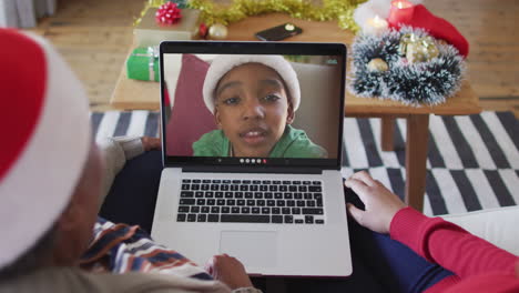 madre y hija afroamericanas usando una computadora portátil para una videollamada de navidad con un niño en la pantalla