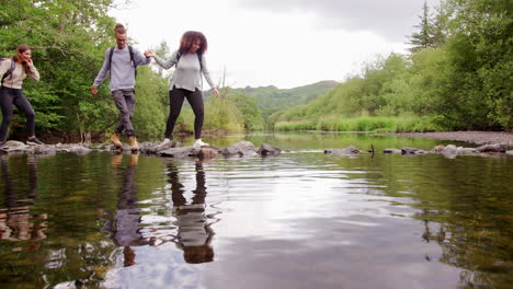 five young adult friends hold hands carefully crossing a stream balancing on stones during a hike, lockdown