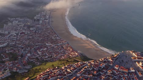 Reveal-shot-of-Nazare-Portugal-with-low-clouds-fog-rolling-over-coastline,-aerial