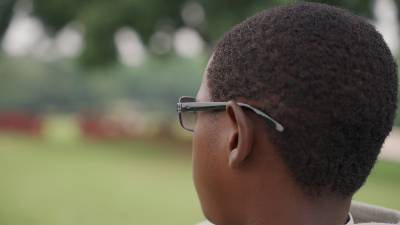 Closeup black son wearing glasses looking over shoulder, soft bokeh park background, short afro haircut, calm afternoon light, candid profile shot conveying introspection, gentle texture of skin