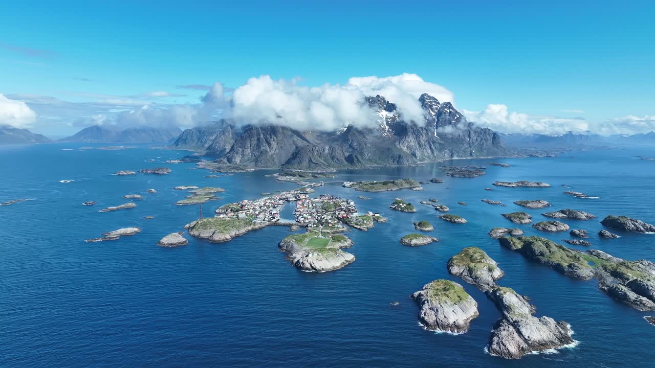 Henningsvaer archipelago and football pitch with sea and cloud capped peaks in Lofoten, aerial approach