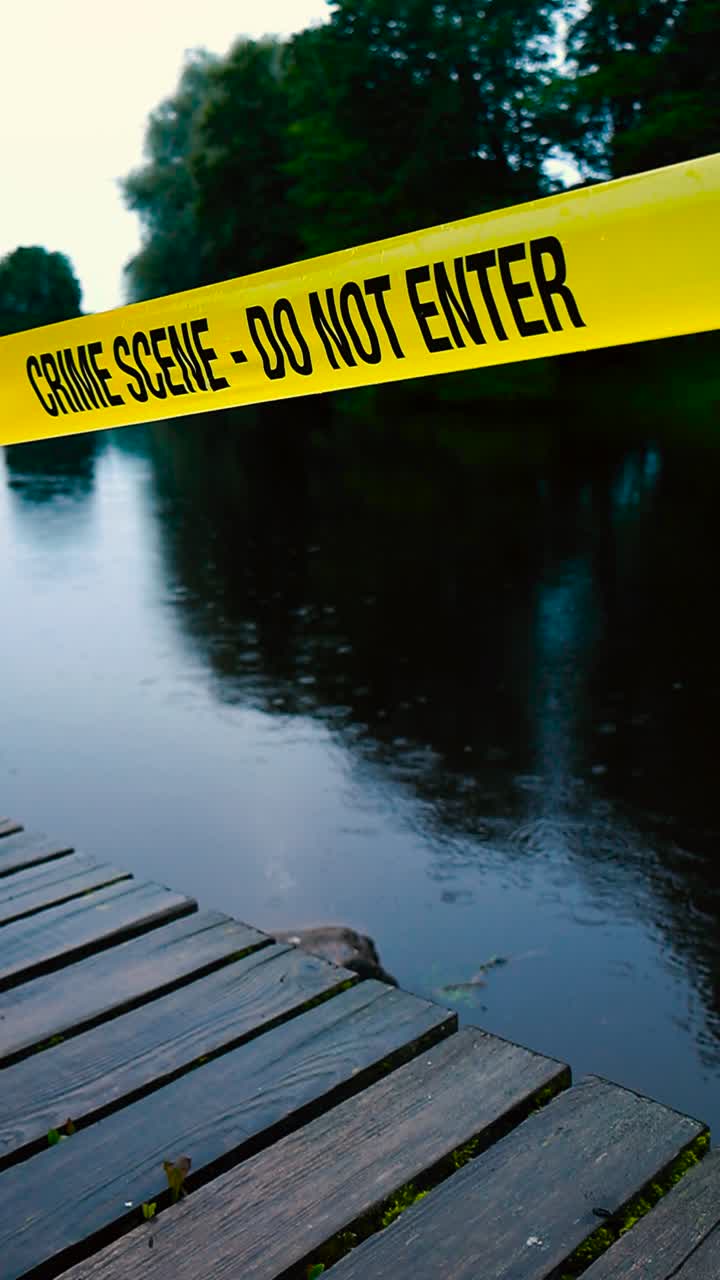 Vertically filmed video of a yellow crime scene Do not enter police tape ribbon pulled above and infront of a dark evening summer lake while it is raining on the water. Green bokeh trees in background