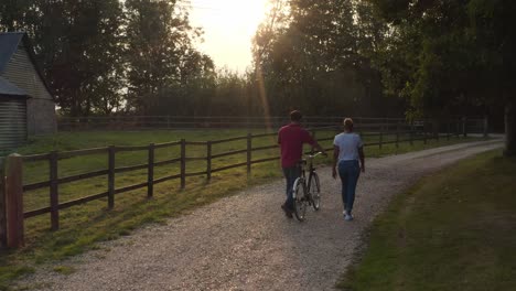 drone rear view shot of romantic couple walking and pushing bike along country lane at sunset