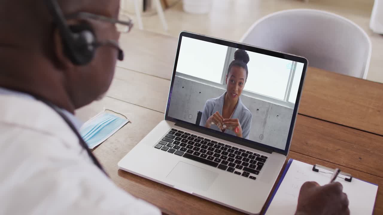 Ein afroamerikanischer Arzt mit Telefon-Headset macht sich Notizen während eines Videoanrufs am Laptop.
