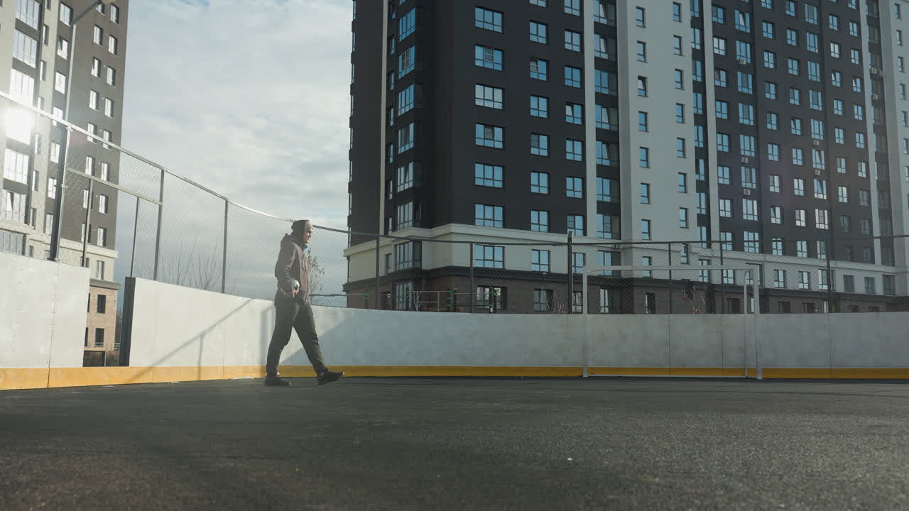 futbolista con capucha y gorra sosteniendo la pelota en la mano derecha pasos en el campo de fútbol con poste de gol, edificio residencial alto, y rayos de luz brillando a través de la escena deportiva urbana