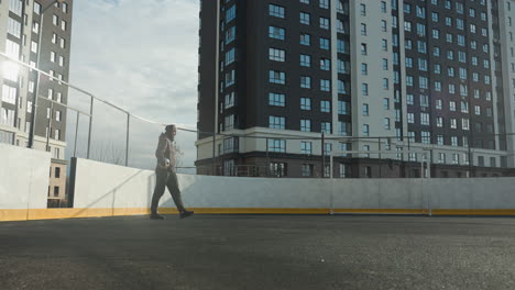 footballer in hoodie and beanie holding ball in right hand steps onto football pitch with goal post, tall residential building, and light rays shining across urban sports scene