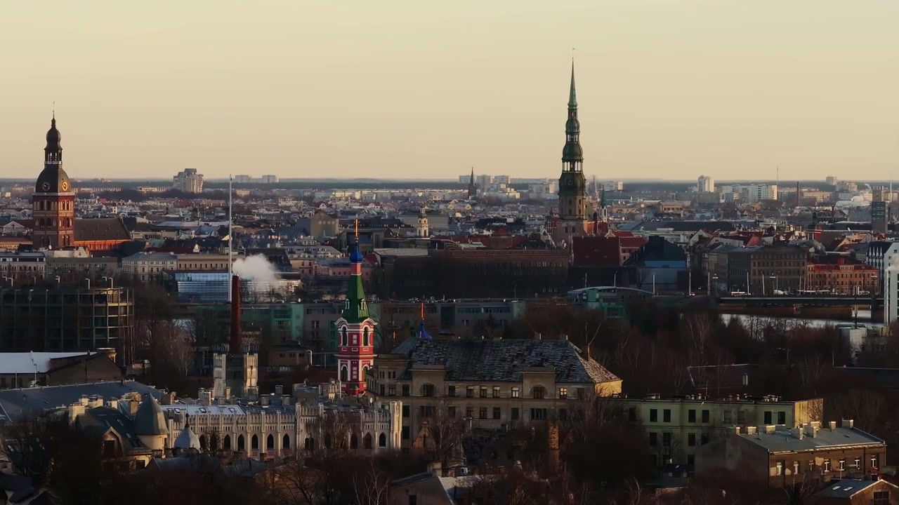 La telecamera gira a destra rivelando la Chiesa della Santissima Trinità, con le guglie della Città Vecchia di Riga sullo sfondo