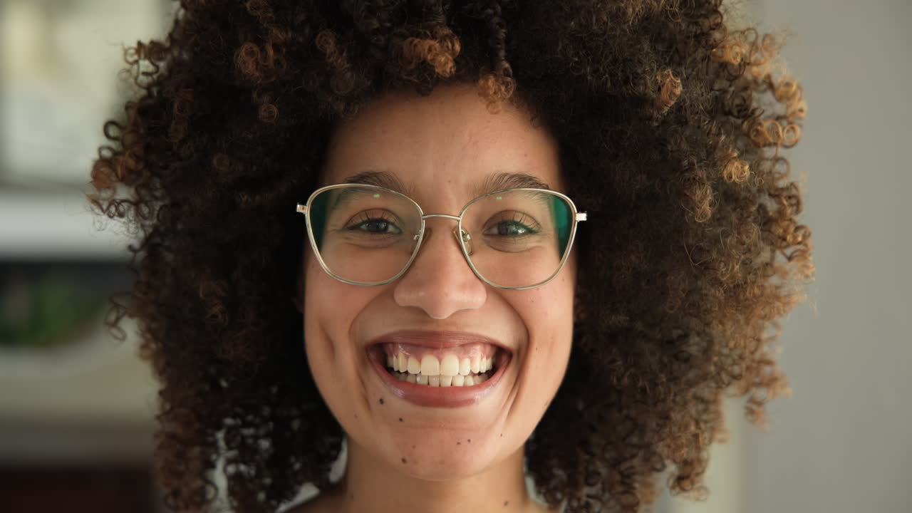Smiling woman with curly hair wearing glasses, expressing joy and confidence, at home