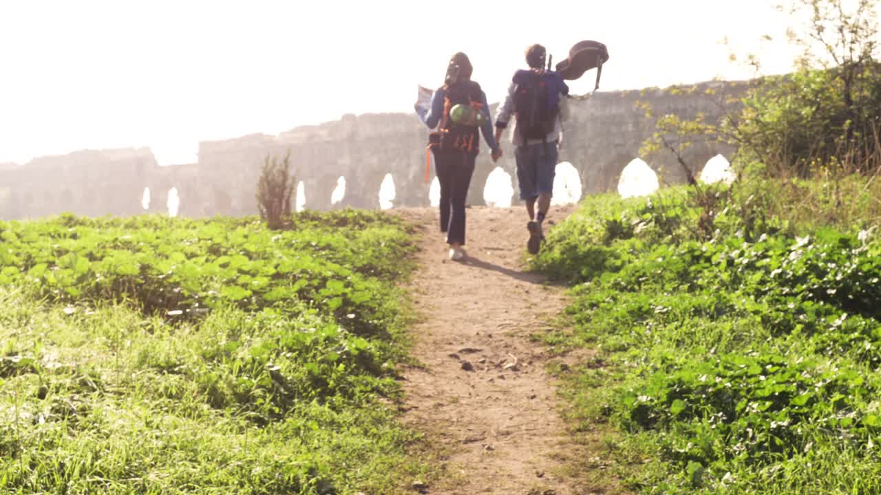 젊은 사랑스러운 커플, 배 여행객, 관광객들이 손을 잡고 로마의 수로를 향해 걸어가고, 파르코 데글리 아키도티 공원, 로마의 유적, 로맨틱한 안개가 가득한 해가 뜨고, 기타와 잠자리 봉투, 느린 동작.