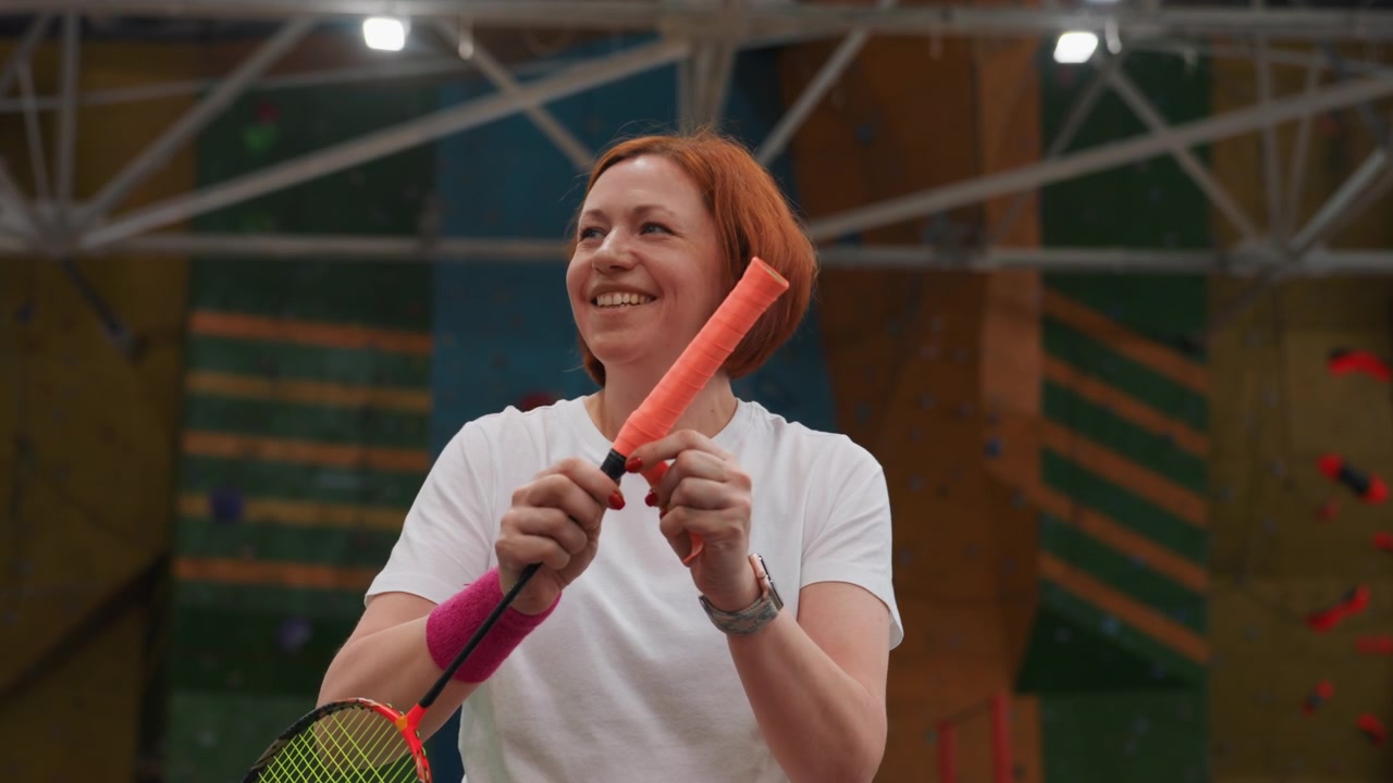 Kvinne undersøker en fjærball ved siden av en badmintonracket i en innendørs gymsal, fokusert uttrykk med armbånd, lys oransje fjærball, hender som justerer fjæren, trenerlignende holdning og treningsøkt.
