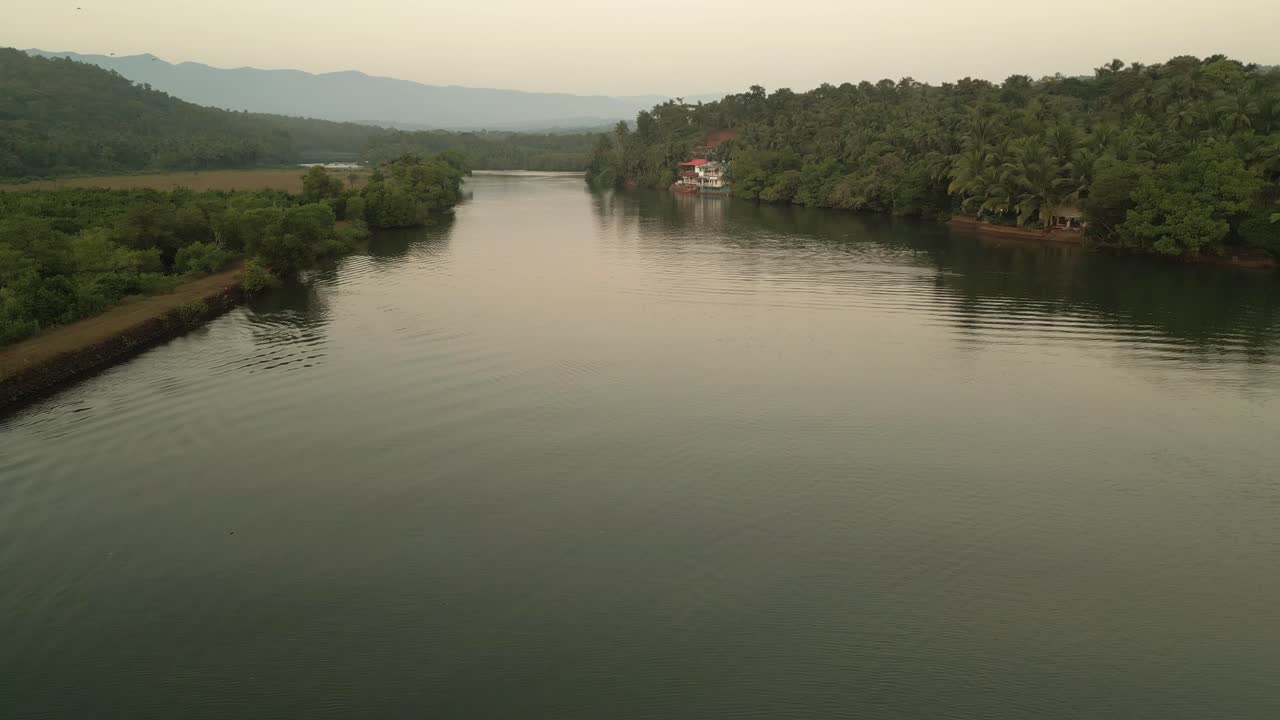 A serene lake flanked by lush green trees on both sides, with majestic mountains rising in the background. The calm water mirrors the vibrant trees Goa India Drone