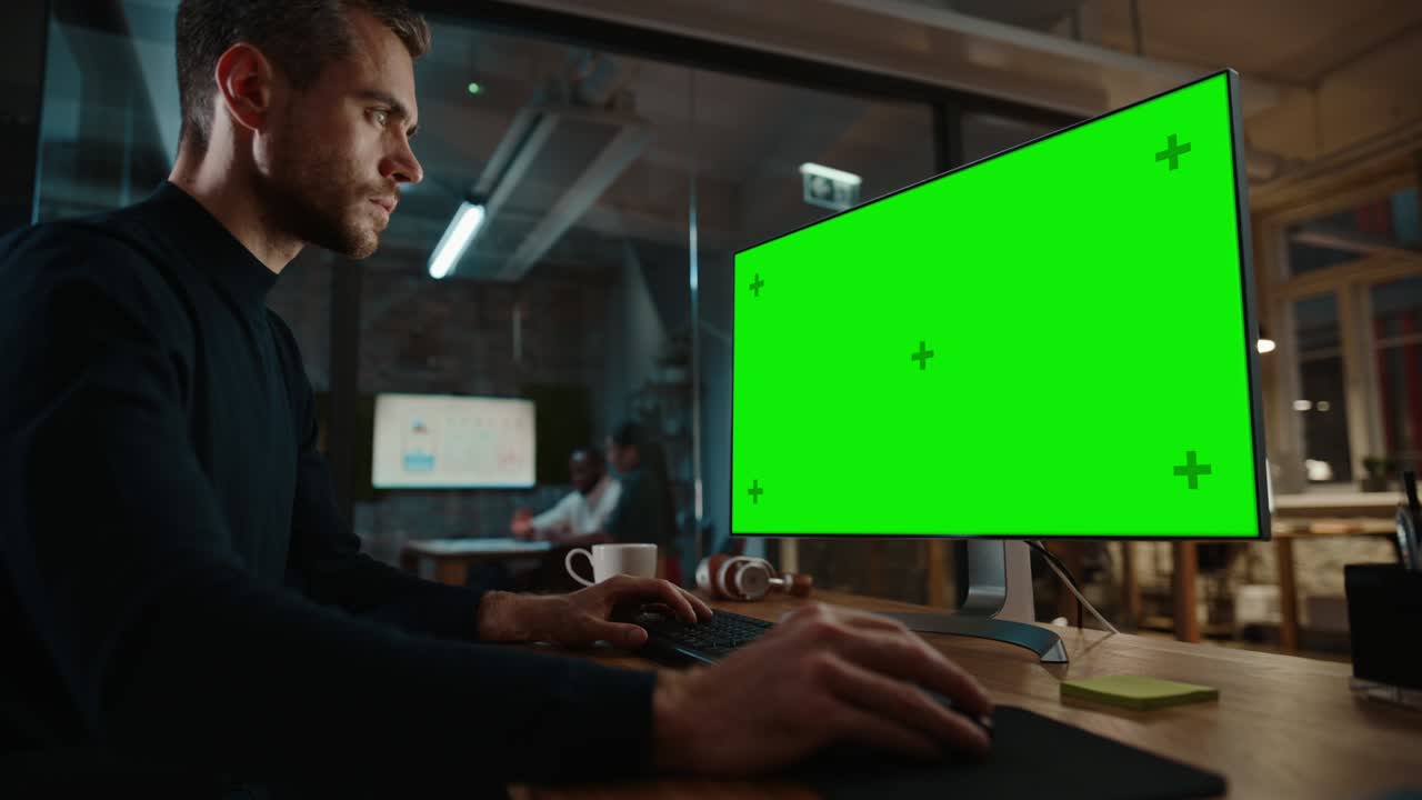 Young Handsome Specialist Working on Desktop Computer with Green Screen Mock Up Display in a Busy Creative Office with Colleagues. Male Manager with Trimmed Beard is Wearing a Casual Black Jumper.