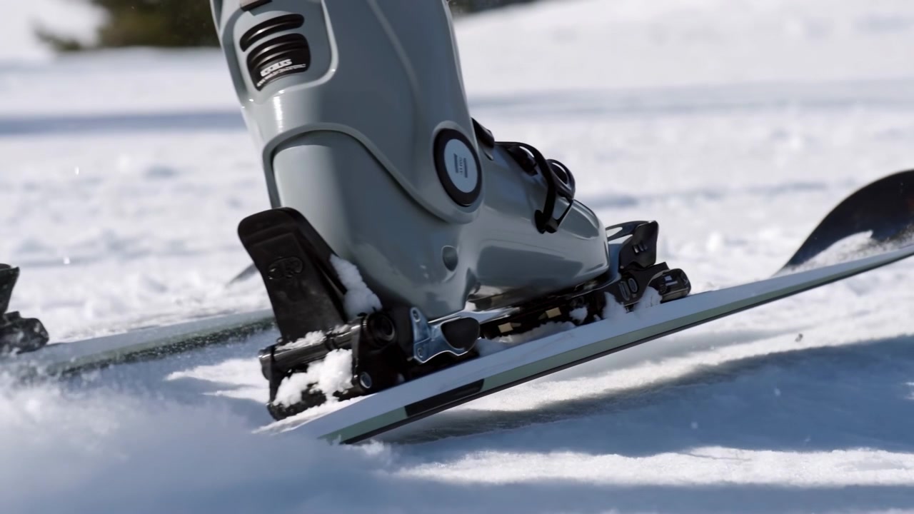 Low-angle motion shows ski boot engaging binding while ski edge lifts from packed snow, sending powder outward with visible pressure release, winter light, cold texture and dynamic alpine movement
