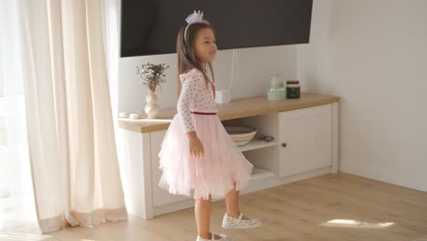 a young girl dances joyfully in a bright, cozy living room. she wears a pink dress with a heart design, moving playfully in the sunlit, minimalist space with neutral decor and soft natural light.