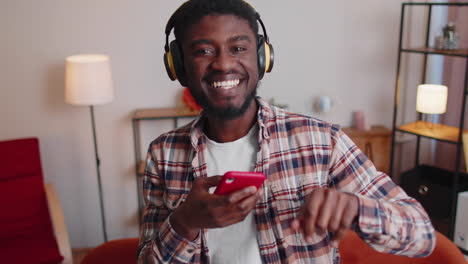 Un-Joven-Muy-Alegre-Con-Auriculares-Inalámbricos-Bailando,-Cantando-En-Un-Acogedor-Sofá-En-La-Sala-De-Estar-De-Casa