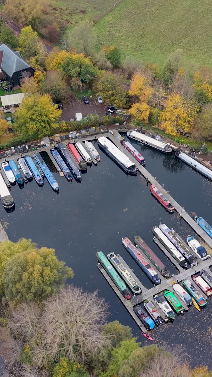 Vertical drone orbits Harlow Marina in Essex. Autumn trees surround the calm, glass-like water, dotted with floating leaves and colourful canal boats, creating a serene and seasonal aerial view