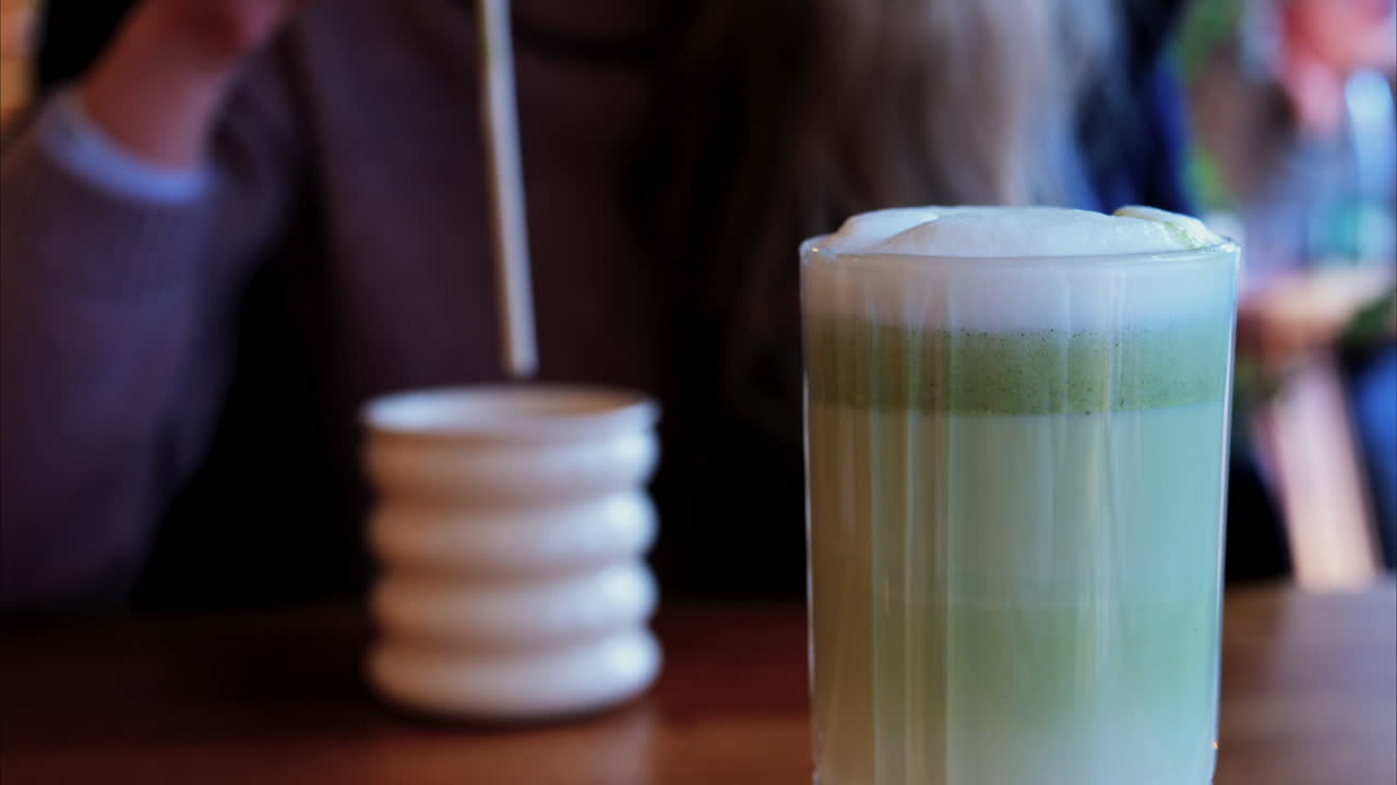 Close up of a matcha latte with a woman drinking at a table at a cafe on the background