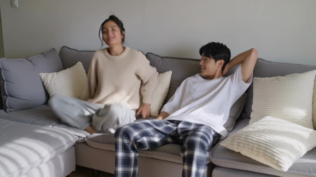 Pasangan mengenakan sweter krem bermain dengan bantal, menyesuaikan posisi berbaring di sofa ruang tamu untuk beristirahat.