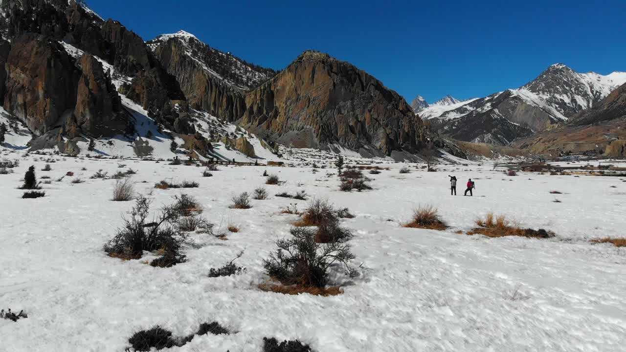 Two people in a snow taking pictures with a big dslr camera and telephoto zoom lens. Sunny day in winter. Annapurna Conservation Area, Manang Valley, Nepal. Aerial Drone shot going forward.