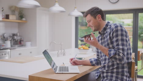 man working from home using laptop on kitchen counter talking into microphone of mobile phone- shot in slow motion