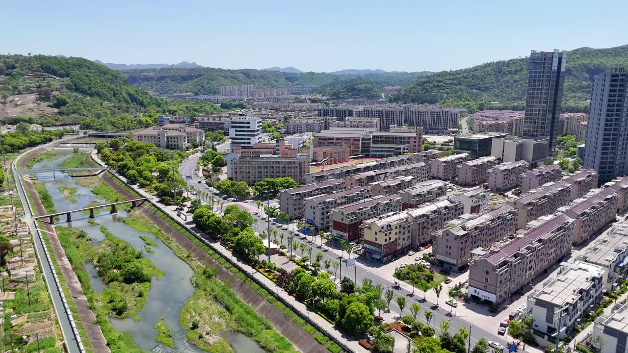 Aerial view of Shaoxing in Zhejiang Province, China, featuring residential blocks, urban buildings, a flowing river, and lush green hills under clear blue skies.