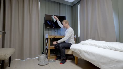 man relaxes on stool fishing through hatch in hotel room floor, cozy indoor lofoten scene