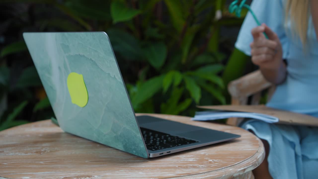 una chica en un vestido azul remotamente en línea trabajando detrás de una computadora portátil y mirando a la pantalla el patio trasero con plantas verdes