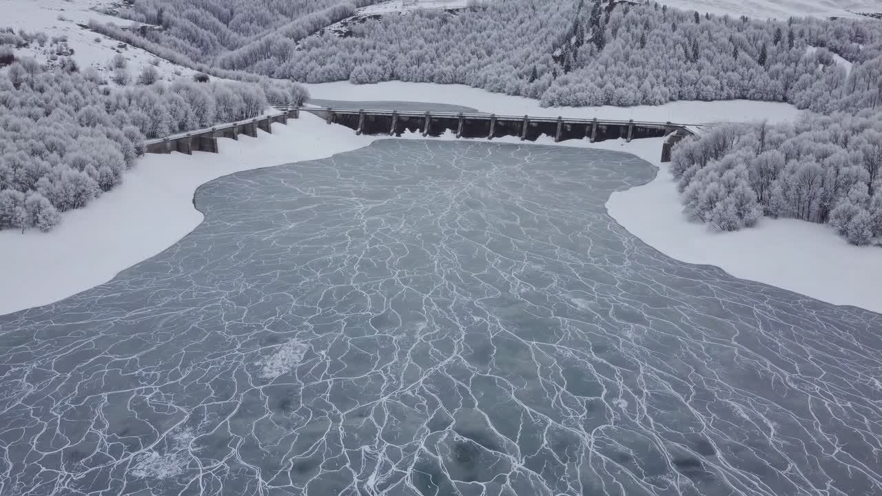 Aerial perspective captures a frozen lake with stunning ice patterns, bordered by snow-laden trees and a dam, illustrating the tranquil essence of winter's landscape