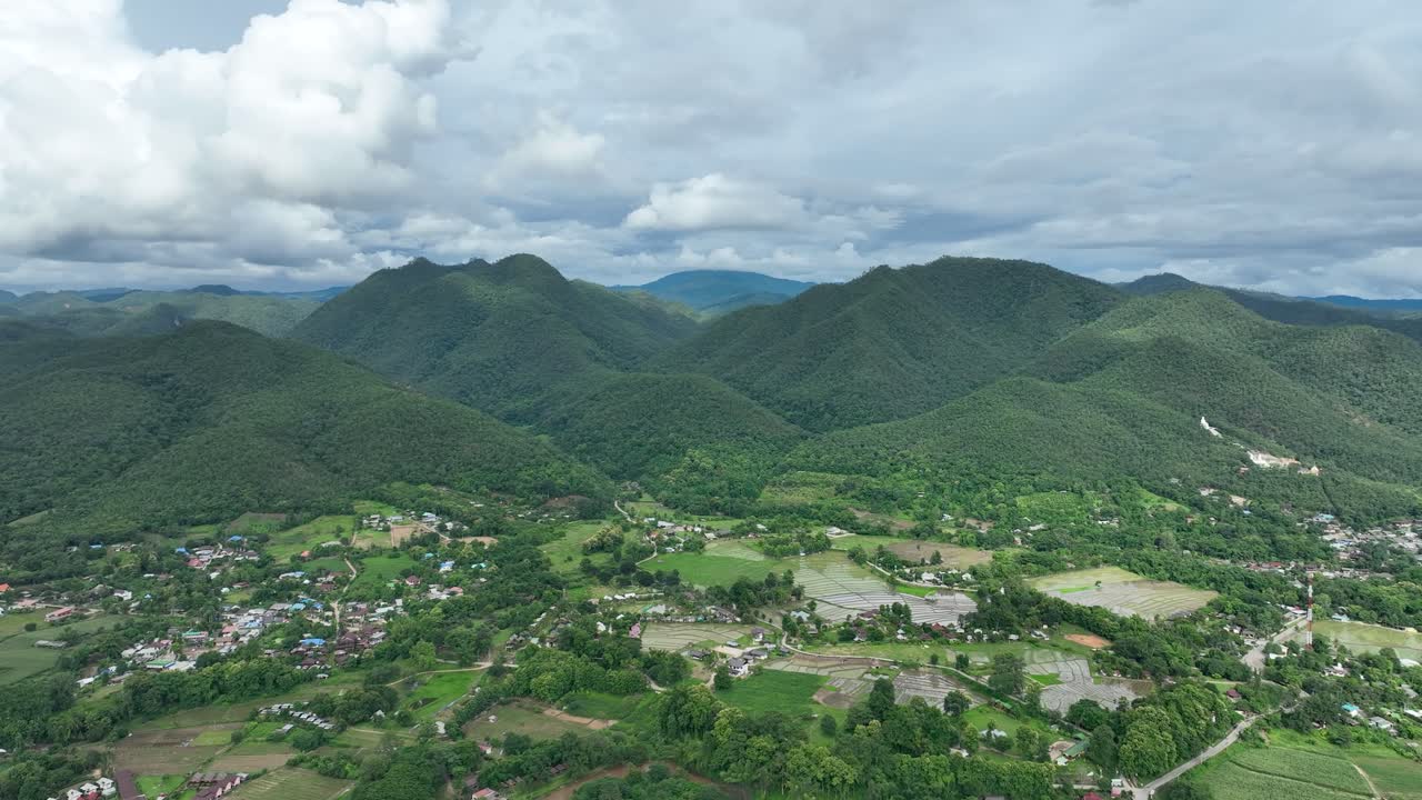 Aerial panorama of rural town Pai in northern Thailand, surrounded by rolling green hills, rice paddies, and tropical vegetation. Peaceful countryside scene under cloudy sky