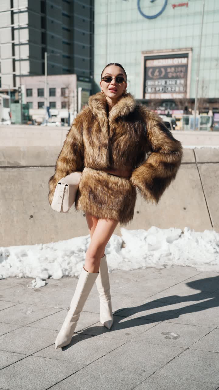 A stylish woman model at DDp posing for photographer in a snowy urban setting, wearing a luxurious brown fur coat, a short skirt, white knee-high boots, and carrying a chic white handbag