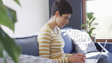 Happy-caucasian-woman-sitting-on-sofa,-using-laptop-and-smiling-in-sunny-house